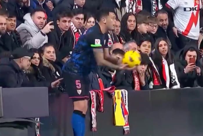 Sevilla's Lucas Ocampos pictured preparing to take a throw-in during a La Liga game against Rayo Vallecano as a fan wearing a baseball cap in the front row appears to poke the player's butt with his index finger
